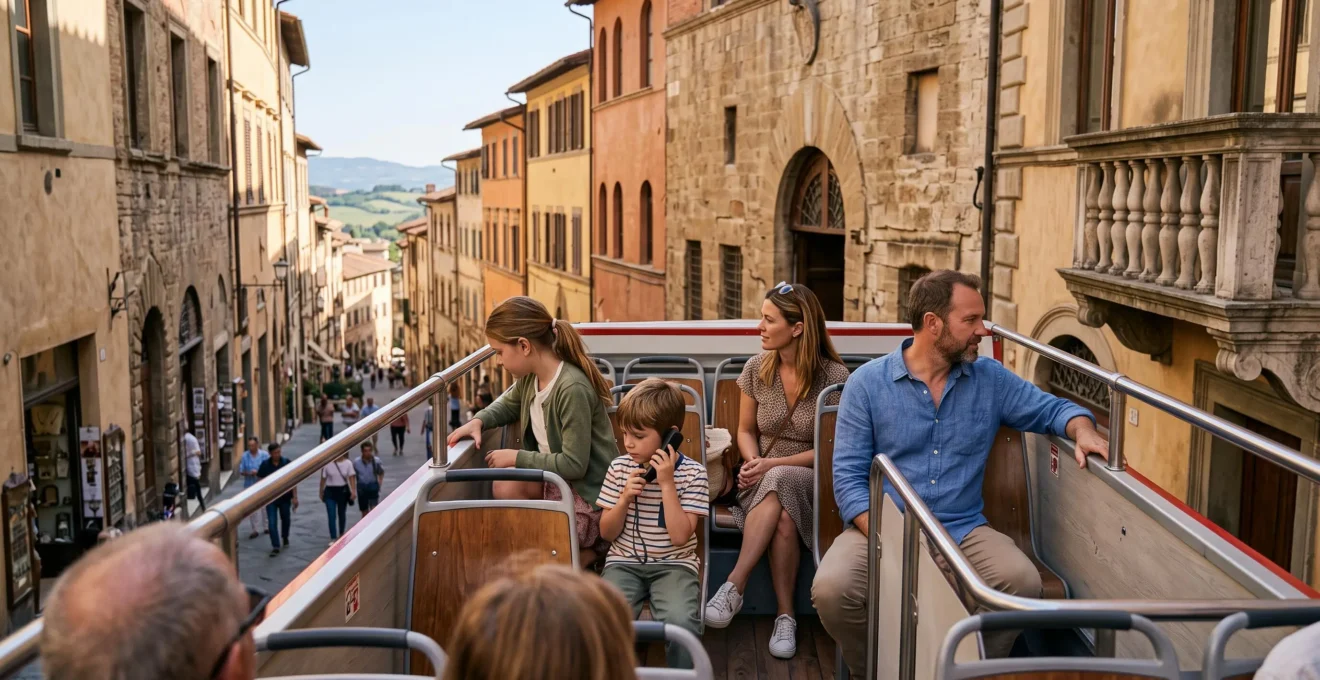 A family with two children seated on the open upper deck of a sightseeing bus traveling through a Tuscan city street.